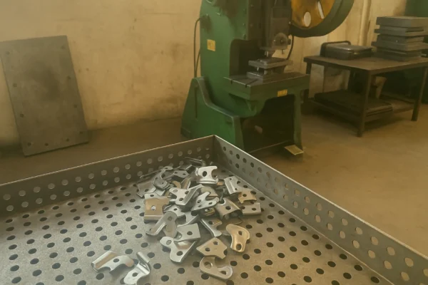 Stamped metal brackets in a perforated parts tray with a stamping press in the background, showing sheet metal stamping production.