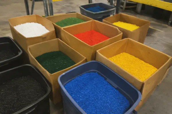 Colorful plastic resins in bins, including red, yellow, blue, green, and black pellets used for injection molding or manufacturing.