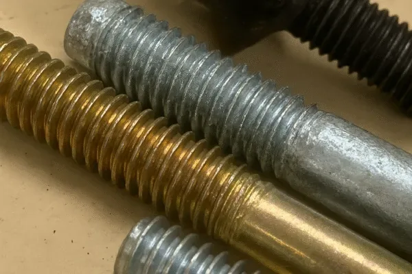 Close-up of threaded metal bolts with different surface finishes, highlighting industrial fastener threads on a workbench.