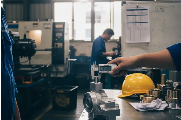 Digital caliper measuring a machined metal component on a shop-floor workbench, with safety helmet and machining equipment in the background.