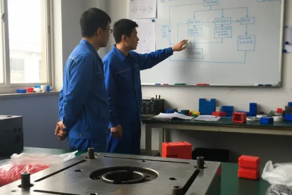 Two engineers in a workshop reviewing a process flow diagram on a whiteboard, with tooling and parts on a workbench in the foreground.