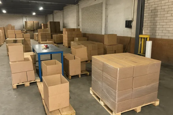 Packaging area in a warehouse with stacked cardboard boxes and a pallet wrapping machine.
