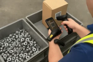 Worker scanning QR code on a carton beside bins of metal components, showing traceability and inventory control for OEM manufacturing.