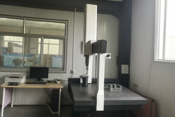 High-resolution image of a coordinate measuring machine (CMM) with granite surface plate, scanning setup for custom metal parts, placed next to a workstation with a computer in a laboratory.