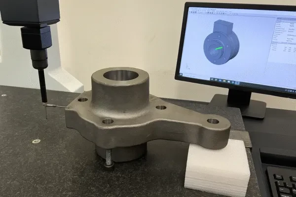 Coordinate measuring machine (CMM) probing a forged and machined hub bracket on a granite inspection table with CAD model on monitor